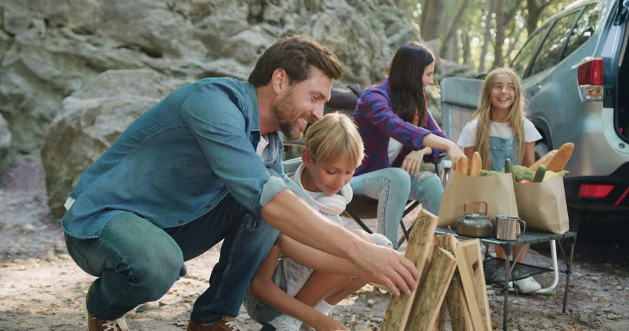 Father and son making together campfire enjoy spend camping evening time outdoors on nature. Smiling mother and daughter are preparing food for a picnic on background. Children with parents are | Shutterstock HD Video #1110610559