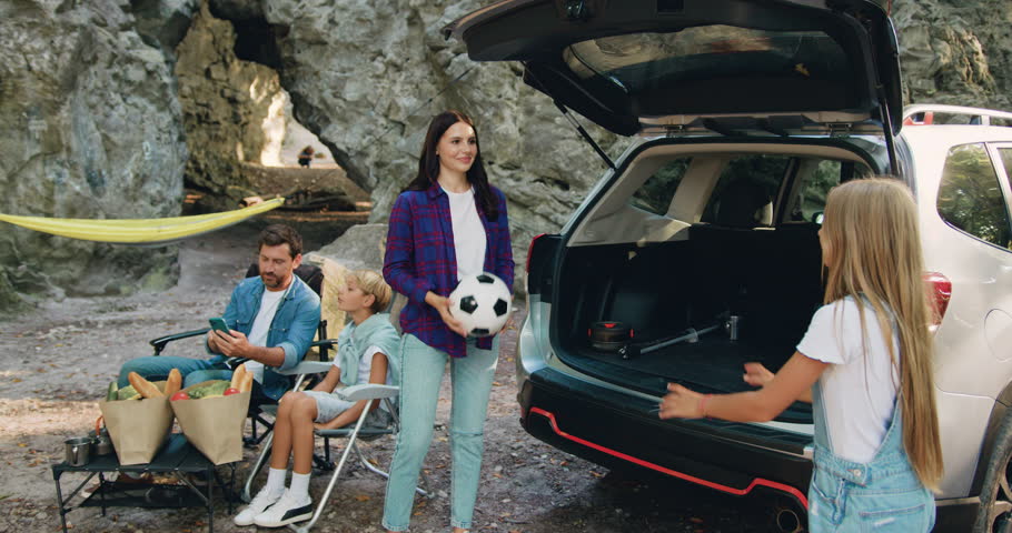 Attractive mother and daughter are playing with ball near car father with son using smartphone during holiday picnic in nature. Children with parent play with ball on grass. Family vacation picnic on | Shutterstock HD Video #1110610569