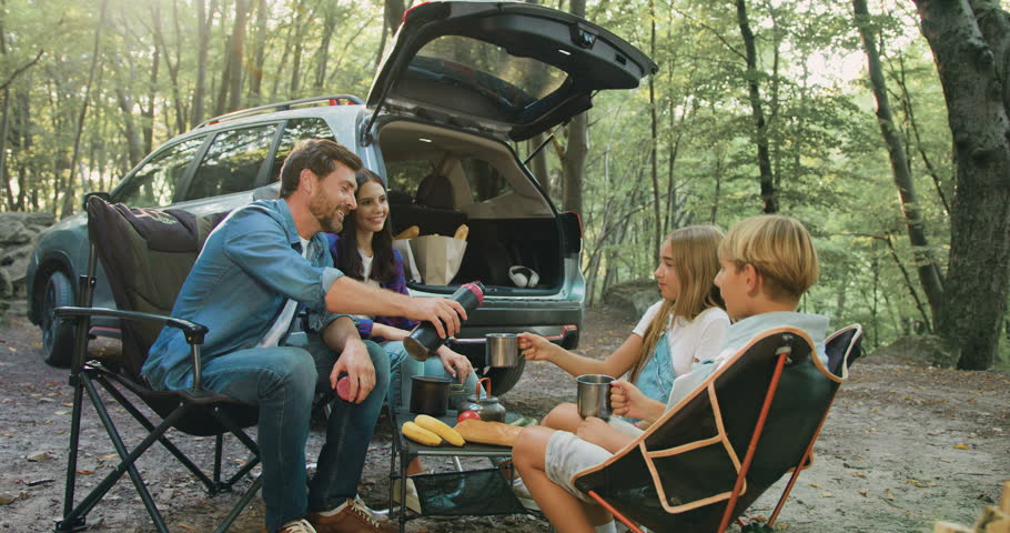 Father mother and children bonding in forest nature, drinking hot tea witch thermos sitting on table near parking car in sunny day. Attractive bearded man pouring hot drink from thermos to pretty kids