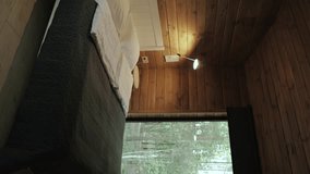 Dolly shot of cozy scandinavian illuminated bedroom with a bedside lamp and white double bed covered by grey sheet cover, wooden wall and view from the window to the forest, slow motion.