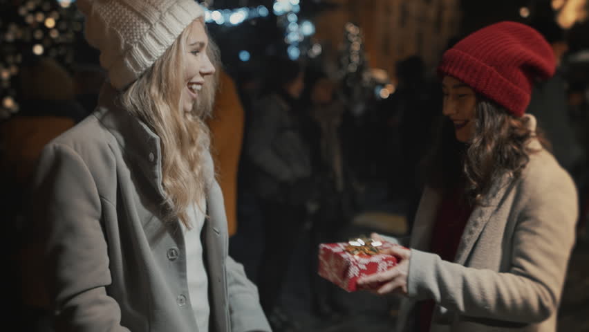 Two women present gifts each other. Happy multiracial friends celebrate Christmas New Year Xmas Eve night together smiling and hug, illuminated background. Young people have joyful time meet at square Royalty-Free Stock Footage #1110611749