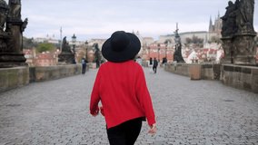 Happy stylish Asian woman in red sweater and hat walking back to camera through Charles bridge. Young tourist traveler visit Prague. Sightseeing in Czech. Smiling girl enjoy holiday vacation abroad