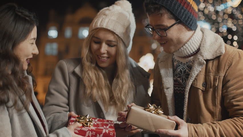 Two women guy present gifts each other. Happy multiracial friends celebrate Christmas New Year Xmas Eve night together smiling hug at illuminated background. Young people have joyful time at square Royalty-Free Stock Footage #1110611829