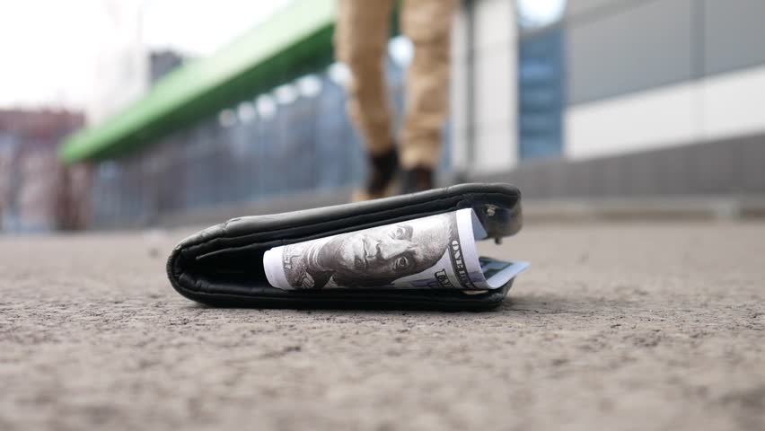 Close-up of a lost wallet on the asphalt with hundred dollar bills sticking out of it and a random person picking it up