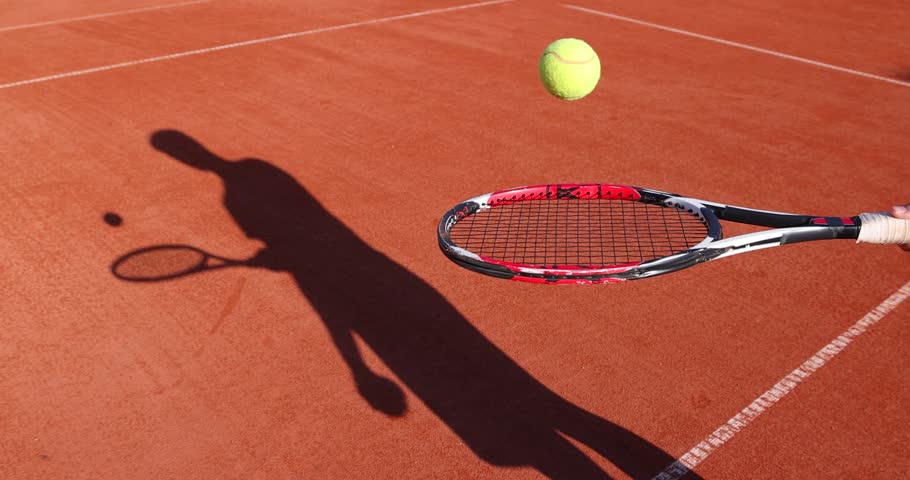 Teenager taps ball on tennis racket. He practices tennis on a clay court, slow motion. His shadow on the sports field