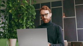 A young red-haired guy, a programmer or entrepreneur in glasses, works in a stylish cafe behind a laptop. Freelancer works remotely. Online communication. Small business. - Powered by Shutterstock - Get 15% off with code: PIKWIZARD15