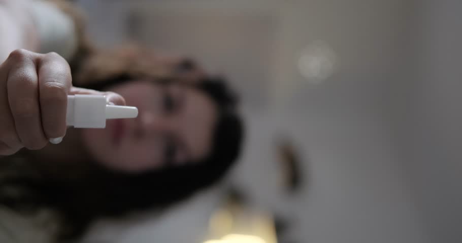 The vertical video in the foreground is spraying nasal spray to treat a runny nose, with a woman's face in the background. Spray drops in the light in slow motion Treatment of runny nose and sinusitis