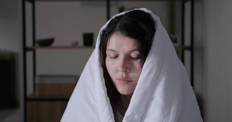 Portrait of a sick young brunette woman who is wrapped in a white blanket and spraying a spray into her nostrils to treat a runny nose. Treatment of a runny nose, nasal drops. Sinusitis treatment.