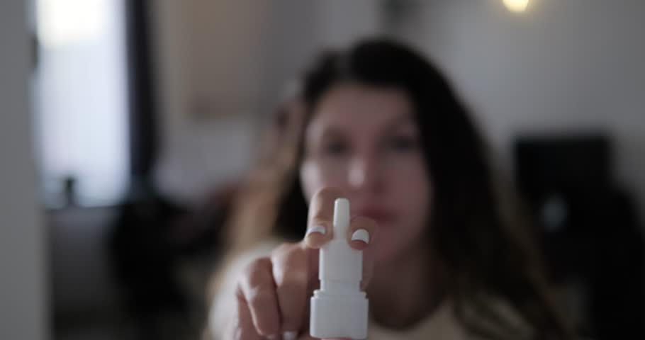In the foreground is spraying nasal spray to treat a runny nose, with a woman's face in the background. Spray drops in the light in slow motion. Treatment of runny nose and sinusitis.