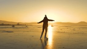 LENS FLARE, SILHOUETTE: A happy woman is skating on a golden shining frozen lake. She is having fun and enjoys dancing on vast empty natural ice rink as the winter sun is setting behind the hills. - Powered by Shutterstock - Get 15% off with code: PIKWIZARD15