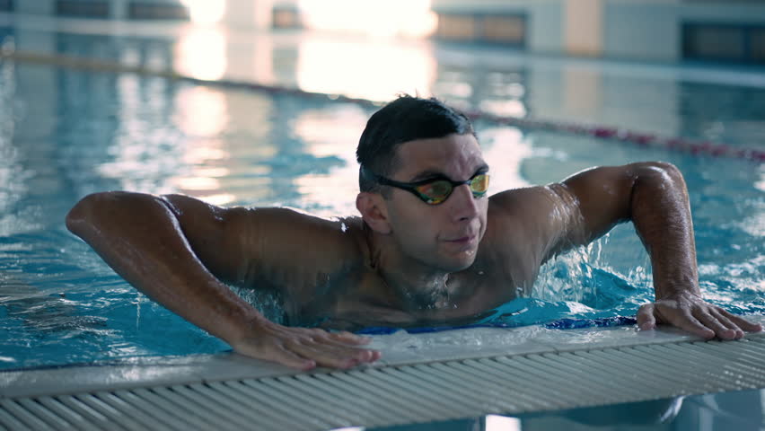 Attractive man with mask jumps out of pool during training. Young athletic swimmer gets on edge finishing exercises in swimpool of sports center
