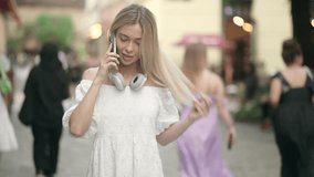 Video of smiling young blonde woman in a white dress talking and laughing with her mobile phone while walking down the street