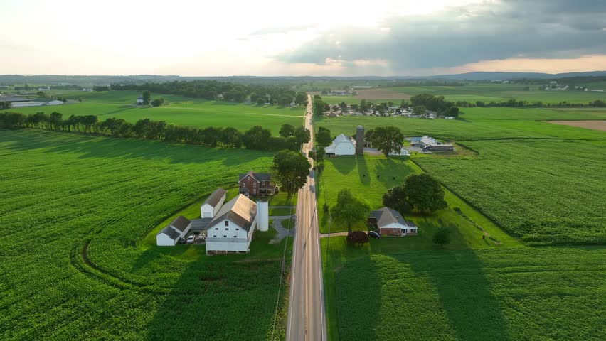 Rural USA. Aerial descending shot above long straight road cutting through green countryside during summer. Farm with fields surrounding.