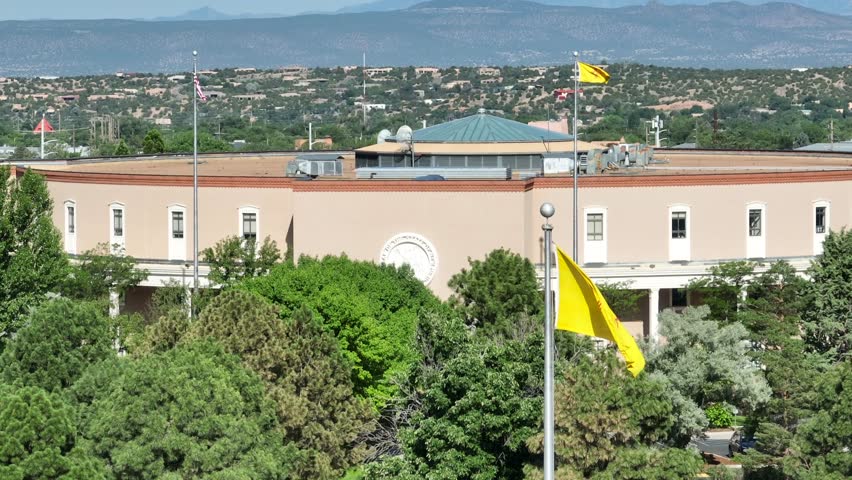 New Mexico state flag waving in front of capitol building in Santa Fe, NM. Aerial establishing shot on bright day.