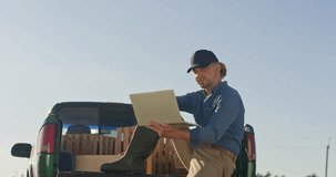Man farmer sitting at pickup truck trunk. Male holding tablet and making video call. Farmer wearing boots an cap making harvest review. Man sitting at trunk with wooden boxes.