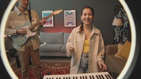 View through ring light of cheerful girl turning on lamp, waving on camera, playing synthesizer and singing as boyfriend playing guitar while going live on social media