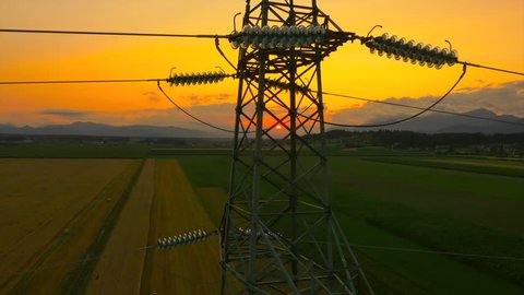 AERIAL: Flying up the high voltage electricity tower and power lines at sunset above the beautiful agricultural field
