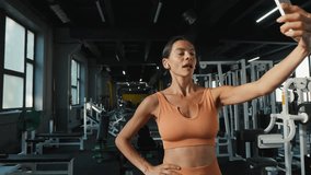 Sportswoman records video showing thumb up gesture and V sign in modern gym. Fitness lady flexes biceps demonstrating well-toned muscles