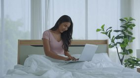 Happy professional Asian businesswoman using laptop sitting on bed at home. Smiling girl sitting, relaxing, video calling, talking about online work or study class. Take notes in a notebook.