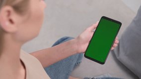 A woman makes a swipe left gesture on a green phone screen while sitting on the couch at home. Swine to the left.