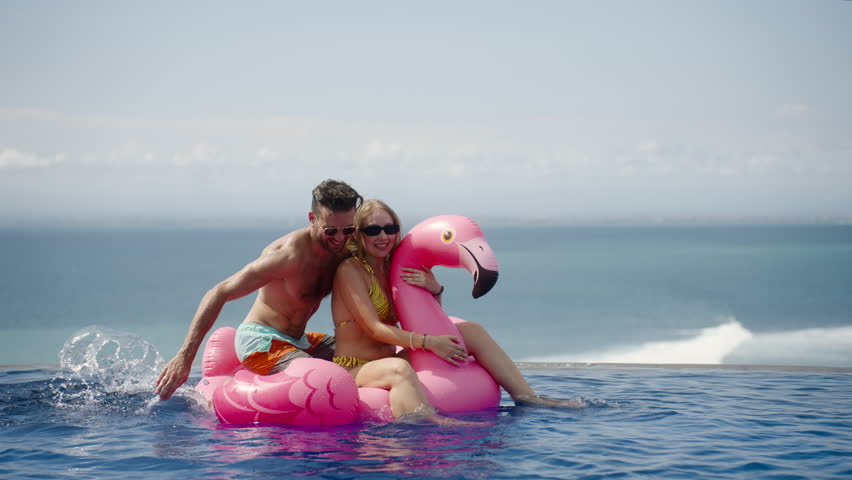 Swimming in pool with pink flamingo. Funny married couple husband and wife having fun on vacation, splashing water emotionally. Honeymoon trip, holiday activity | Shutterstock HD Video #1110651657
