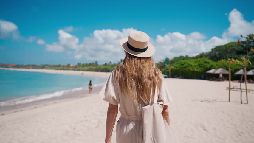 Romantic blonde woman tourist in straw hat walking to ocean sandy beach in sunny day, rear view. Female lady enjoying nature on vacations summer holidays. Travel, tourism, journey, rest relax concept.