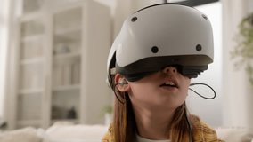 Surprised amazed Caucasian girl kid child schoolgirl playing online video game in 3D world metaverse using virtual reality helmet at home play gaming controller explore cyberspace wearing VR glasses