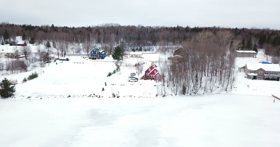 Aerial Panning Shot Of Snow Covered Houses Amidst Trees Near Frozen Lake Against Sky - Houghton, Michigan