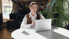 Focused female smiling worker sitting at restaurant using wireless laptop for video call. CEO manager lady in white blouse having distance chat with partners drink coffee during online meeting.