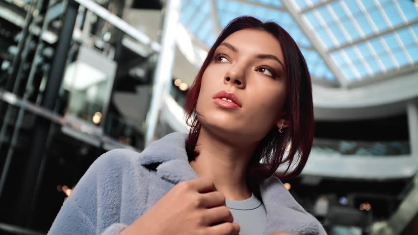 Beautiful fashion brunette woman model posing in gray fur coat clothes presentation at modern shopping mall. Luxury glamour female wearing warm seasonal clothing outfit designer performance closeup