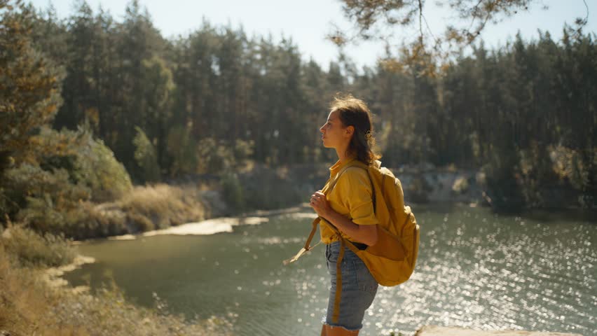 tracking shot Woman hiker walking trail around mountain lake at sunny morning in autumn pine forest at warm day. female traveler with yellow backpack hiking in woods, adventure outdoor lifestyle.
