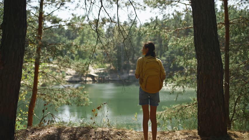 Woman hiker walking trail around mountain lake at sunny morning in autumn pine forest at warm day. female traveler with yellow backpack hiking in woods, adventure outdoor lifestyle.