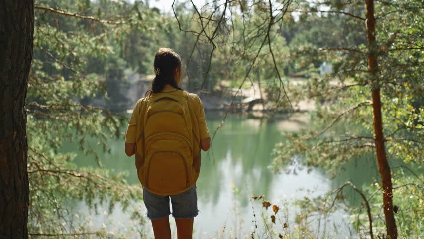 Woman hiker walking trail around mountain lake at sunny morning in autumn pine forest at warm day. female traveler with yellow backpack hiking in woods, adventure outdoor lifestyle.