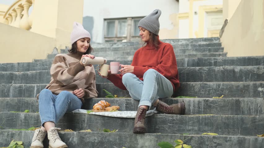 In autumn, two women drink tea from a thermos and eat croissants on the steps of the stairs. Friends in warm autumn clothes chatting outdoors