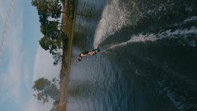 Vertical video. Male wakeboarder riding board with rope jumping slide and smile at tropical water lake aerial view. FPV drone shot man extreme sport trick wakeboarding track at sunny sunlight splashes