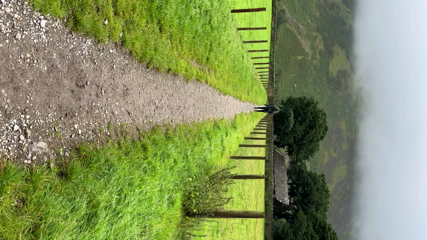 Personal perspective POV following unrecognizable tourist woman, walking gravel footpath between stone walls of sheep pasture in the mist covered mountain Wasdale valley in Lake District National Park