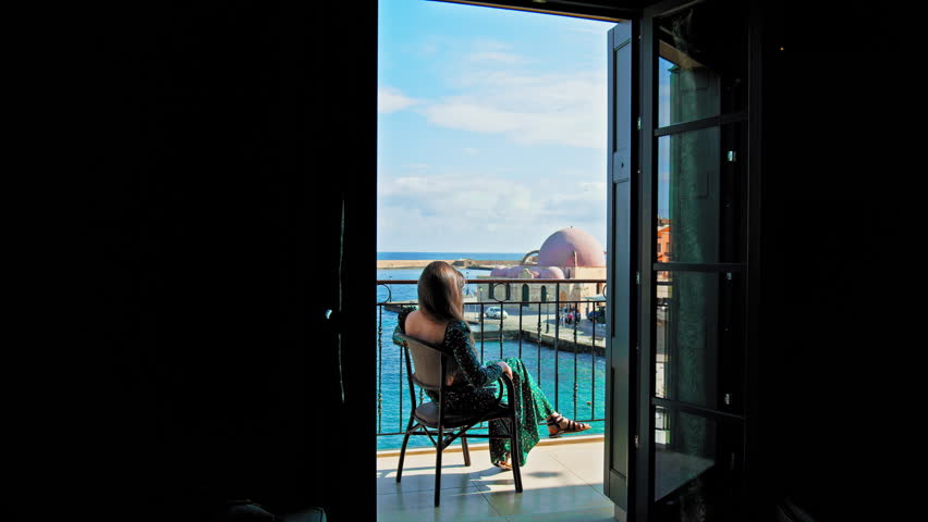 A beautiful girl in a dress enjoying the view from the hotel balcony in Chania, Crete. Young woman overlooking colourful Old Venetian Port of Chania surrounded by turquoise-blue water, mosque landmark