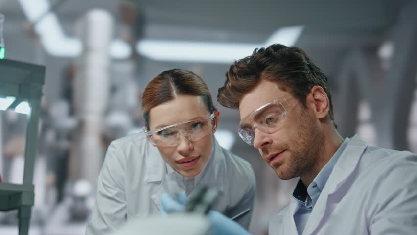 Two chemists examining test tube with experimental medicines in advanced laboratory close up. Woman scientist in glasses showing sample to colleague. Serious researchers in robe working modern clinic.
