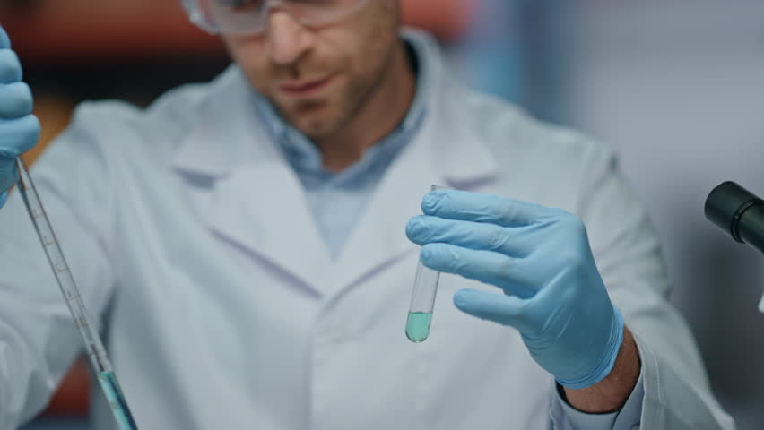 Focused researcher using pipette filling test tube in laboratory close up. Professional man scientist examining medical preparation in clinic. Serious microbiologist in safety glasses working at lab.