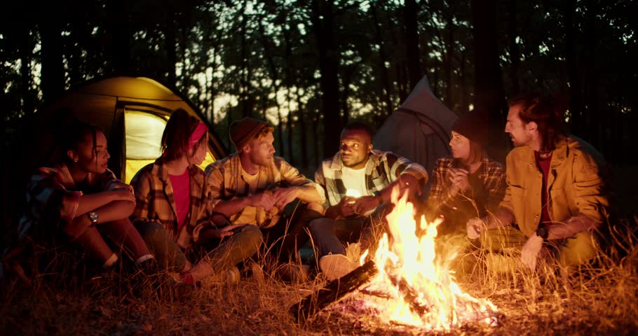 A group of scouts during a rest stop tell each other scary stories near a fire in the night forest with tents in the background
