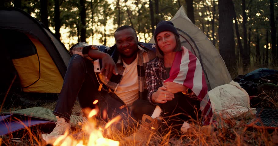 A man with black skin and a girl with a bob in a black hat sit near a fire during a camping stop and hug under the US flag against the backdrop of a beautiful forest and tents
