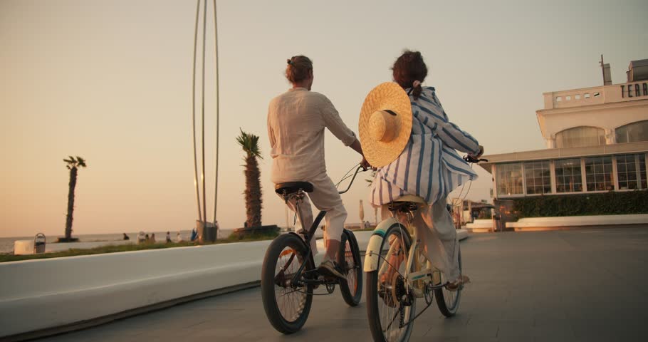 Rear view of a happy guy in white clothes on a Black bicycle and a girl with a Straw hat on a bicycle ride along the beach near the sea at sunrise in summer. Active leisure and date on the go on