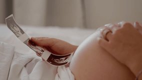 Close up shot side view of unrecognizable pregnant woman admiring sonography picture of her unborn baby and caressing belly, sitting at home. Enjoying pregnancy female touching belly. - Powered by Shutterstock - Get 15% off with code: PIKWIZARD15