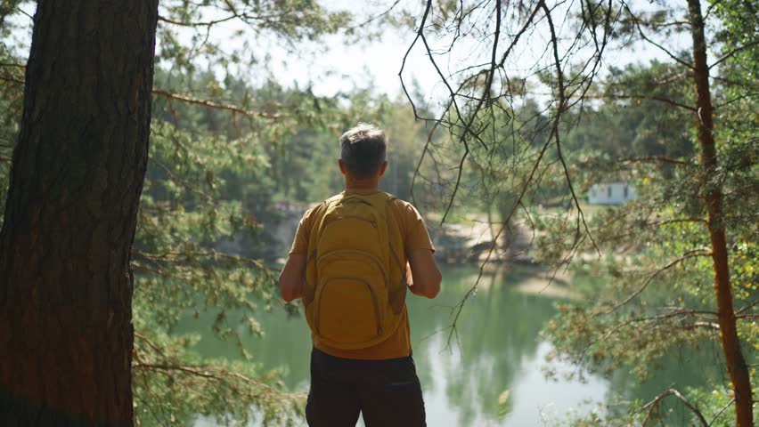 man hiker walking trail around mountain lake at sunny morning in autumn pine forest at warm day. male traveler with yellow backpack hiking in woods, adventure outdoor lifestyle.