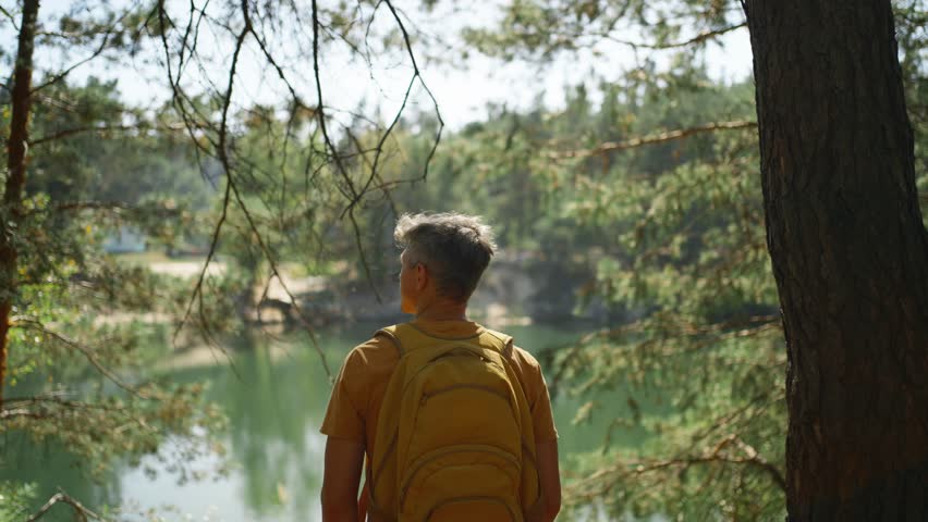 man hiker walking trail around mountain lake at sunny morning in autumn pine forest at warm day. male traveler with yellow backpack hiking in woods, adventure outdoor lifestyle.