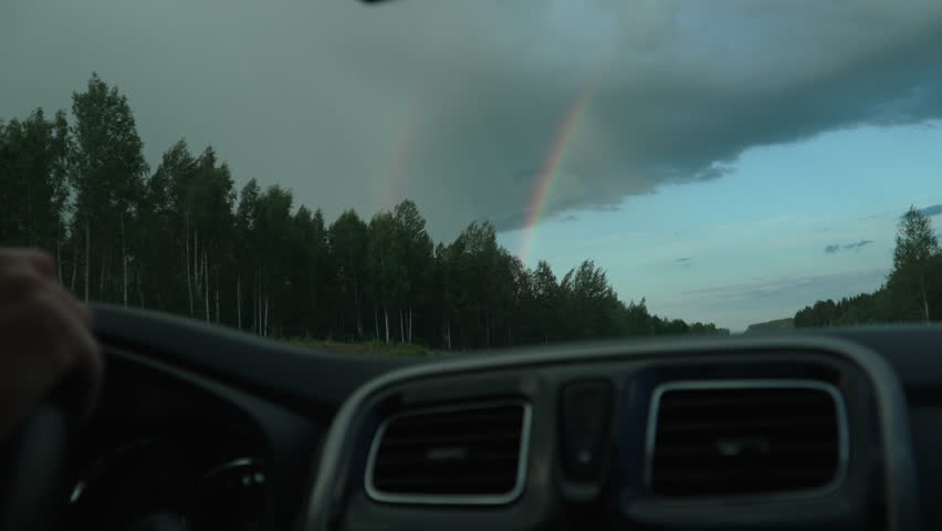 View from inside car to edge of cloud and double rainbow behind forest on highway. Traveling along highway among trees after rain. Air ducts and drivers hands on steering wheel.