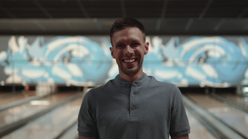 Middle aged caucasian man with hard shadows on his face smiles in bowling club, applauds and shows thumbs up. Happy player in front of bowling alley under light source after successful throw.