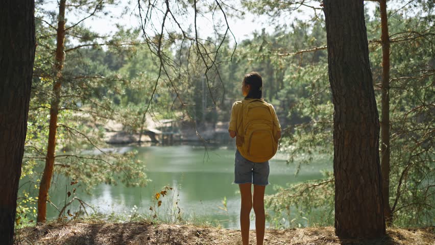 Woman hiker walking trail around mountain lake at sunny morning in autumn pine forest at warm day. female traveler with yellow backpack hiking in woods, adventure outdoor lifestyle.