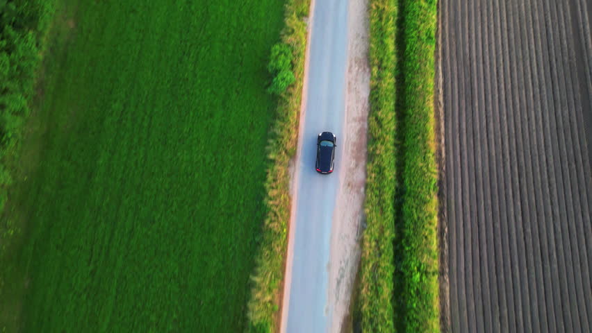 Hovering above, the camera offers a dynamic bird's-eye view: a lone car, a speck of modernity, journeying down a road that cuts through an expansive sea of green. The lush field, a testament to nature Royalty-Free Stock Footage #1110718019