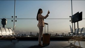 Full Body Back View Of Asian Businesswoman With Rolling Suitcase In Boarding Lounge At The Airport, Waving Hand Having Video Call On Smartphone, Airplane Takes Off Outside The Window